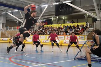 Un attaquant des Geneva Dragons est en extension, sur le point de tirer depuis l'aile droite, face à trois défenseurs et une défenseuse des Val-de-Ruz Flyers, sous les yeux de l'arbitre, accroupi à côté du cadre. En arrière-plan, on voit le public d'une centaine de personnes qui remplit les gradins.