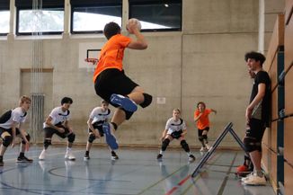 Un ailier droit, de l'équipe Lancy Sharks, en orange, est en extension avec la balle à la main, prêt à faire la passe à l'ailier gauche en fasse. En défense se trouvent une joueuse et 3 joueurs de l'équipe de Fribourg.