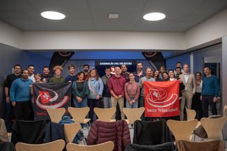 Les participantes et participants (26 personnes) à l'Assemblée des délégués 2024 posent dans la salle où a eu lieu l'assemblée. Ils portent deux drapeaux carrés, un rouge et un noir, chacun avec le logo de Swiss Tchoukball.